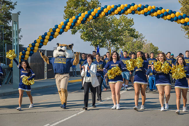 Enthusiastic cheerleaders and bobcat mascot parade under a blue-and-gold balloon arch.